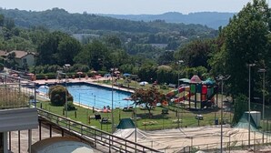 Área de piscina com escorregador, cercada por vegetação e vista distante de colinas.