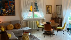 Een moderne woonkamer met een grijze bank, gele kussens, een kamerkleed met een patroon, een eethoek met een ronde tafel en stoelen, en een groot raam met gordijnen.