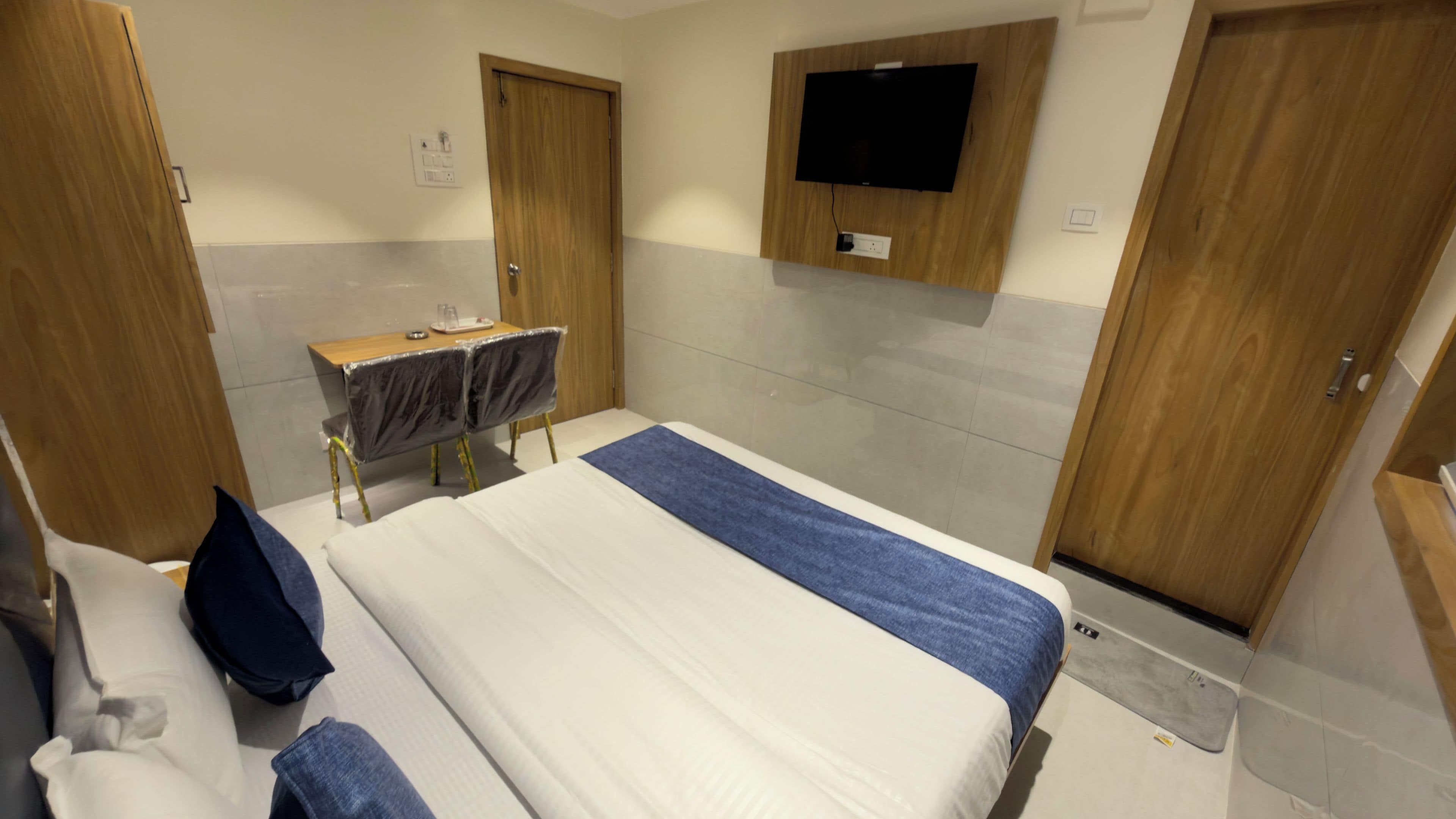 Une chambre d’hôtel comprenant un lit, un bureau avec une chaise, un téléviseur fixé au mur et deux portes en bois.