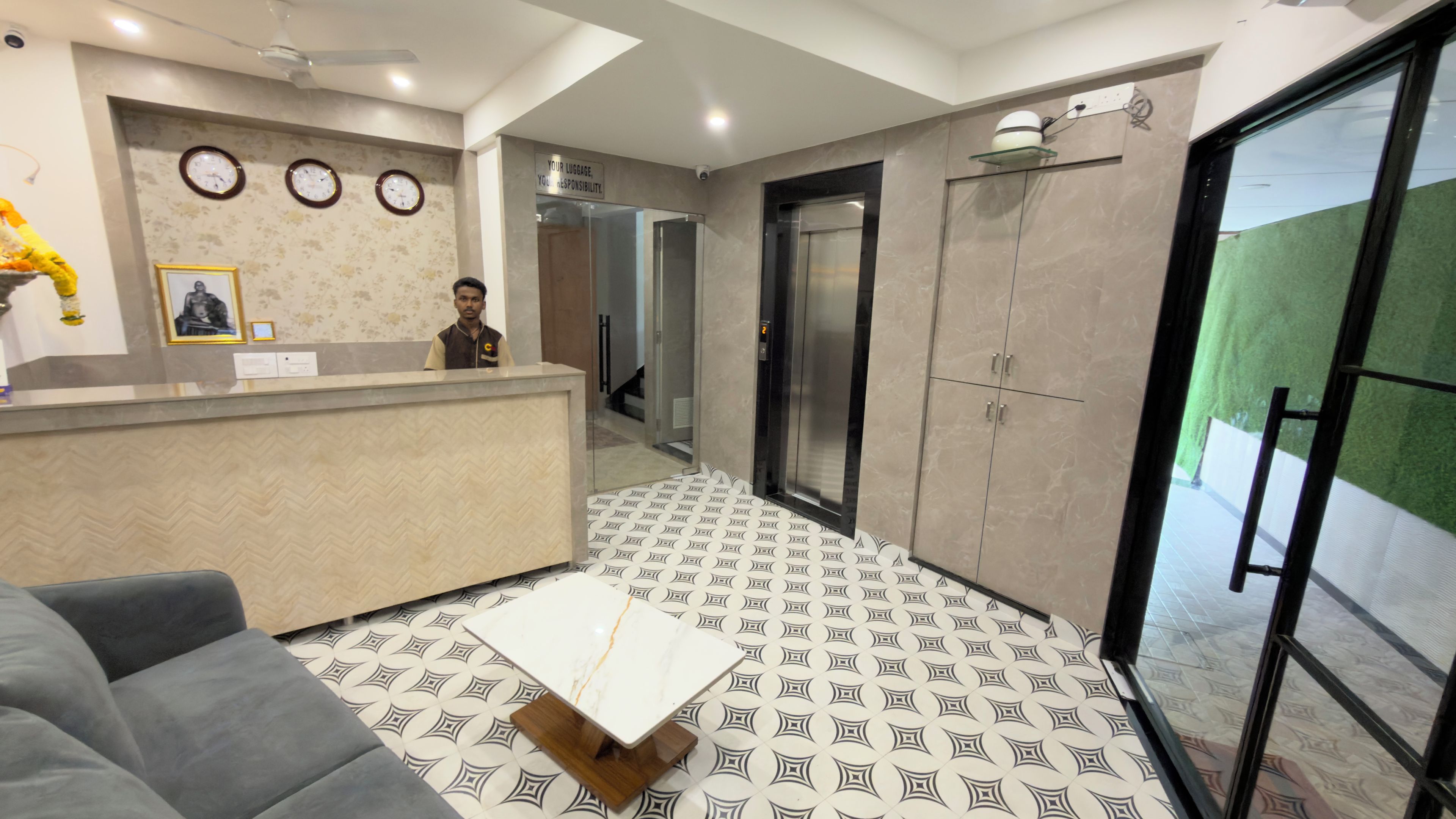 Zona de recepción de un hotel con mostrador, un sofá, suelo estampado y una puerta que da a un ascensor.