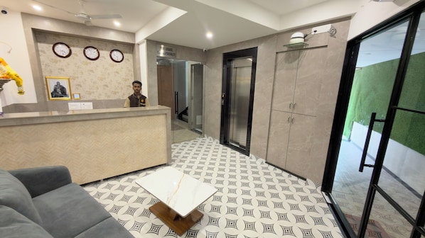 A hotel reception area with a reception desk, a sofa, patterned flooring, and a door leading to an elevator.