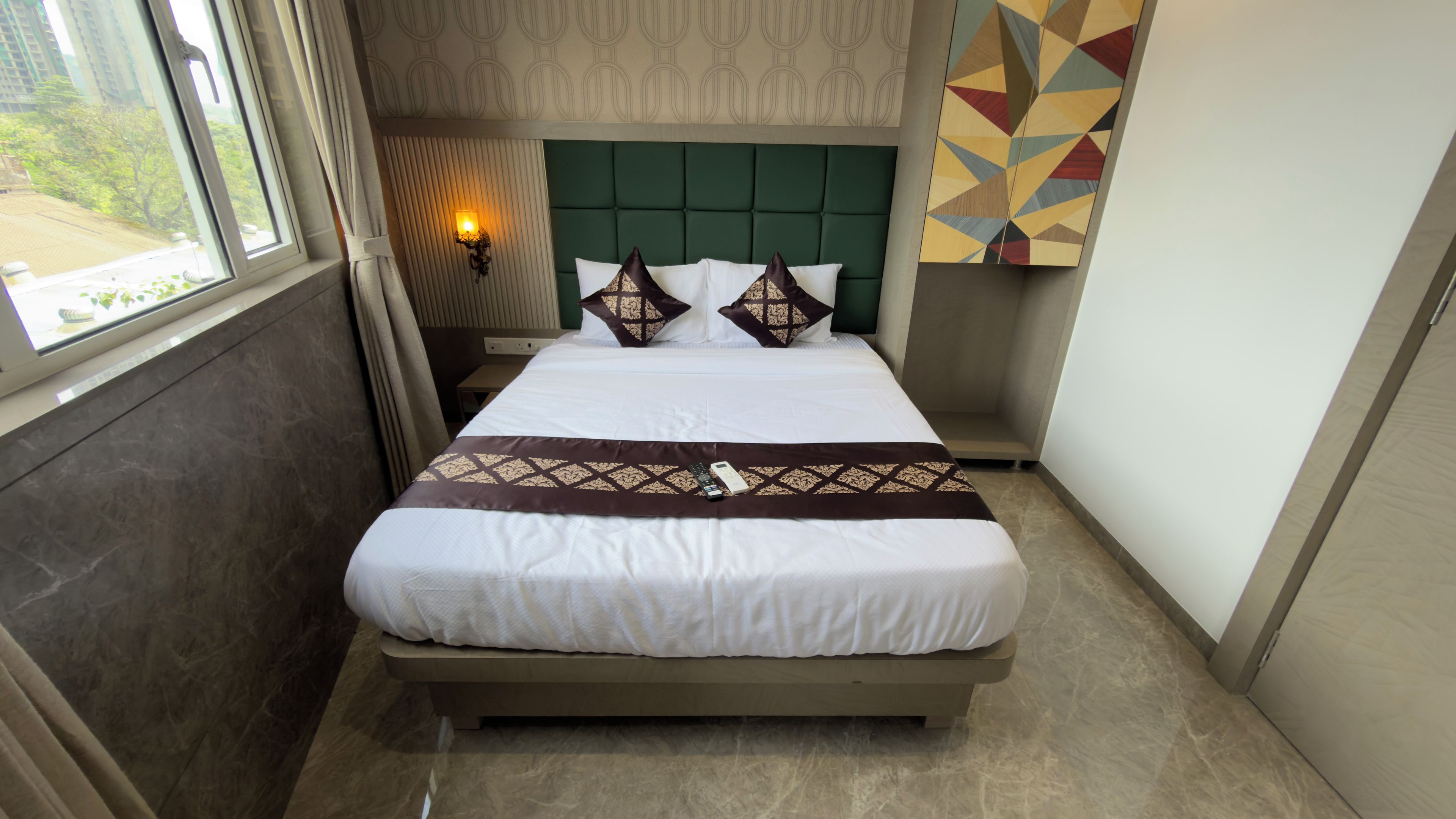 Una habitación de hotel con cama, mesita de noche, lámpara de pared y ventana con cortinas.