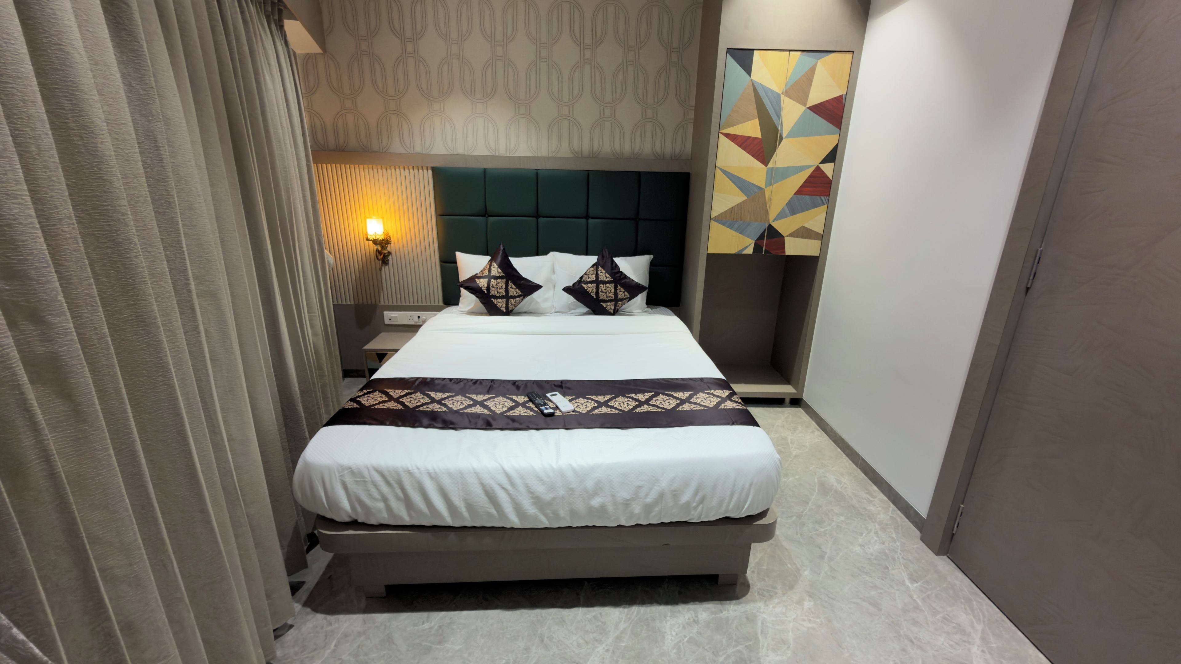 A hotel room with a bed, a bedside table, a wall-mounted lamp, and a geometric wall art piece.