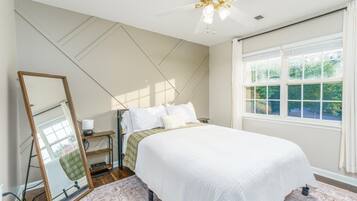 A bedroom with a bed, a mirror, a bedside table, a lamp, and a window with a view of greenery.