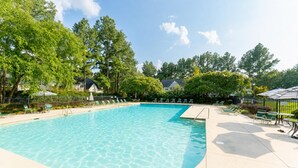 A clear swimming pool surrounded by lounge chairs and umbrellas, with trees and a building in the background. - Garden by the University (Charlotte)