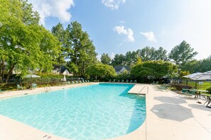 Ein klares Schwimmbecken, umgeben von Liegestühlen und Sonnenschirmen, mit Bäumen und einem Gebäude im Hintergrund.