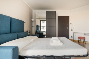 Una habitació d'hotel moderna amb un llit gran, un sofà blau, una taula de treball amb una cadira i una vista de la ciutat per la finestra.