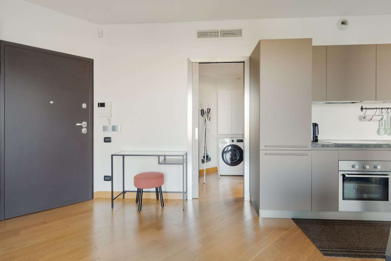 Eine moderne Küche mit Waschmaschine, ein Essbereich mit kleinem Tisch und einem pinken Hocker sowie eine dunkle Tür.