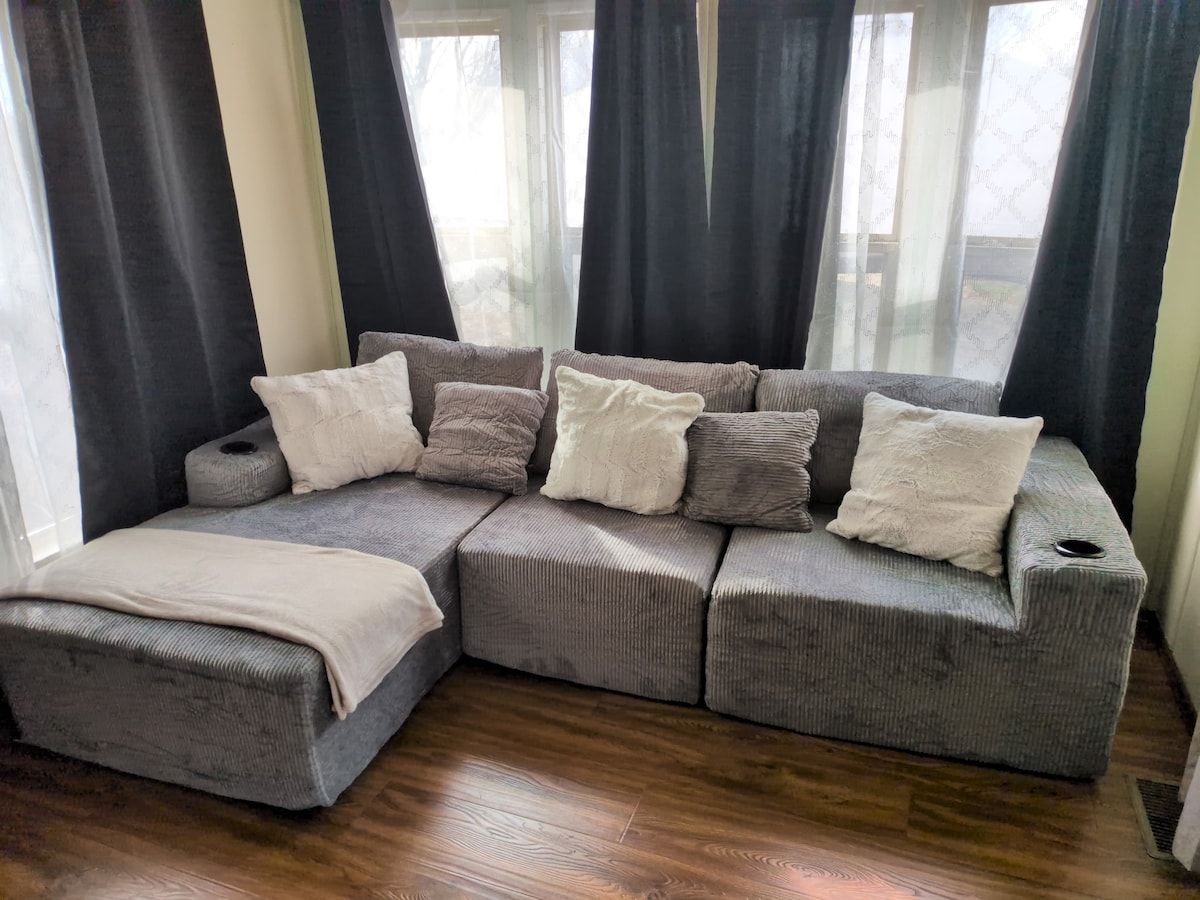 Ein modulares Sofa mit vielen Kissen in einem Raum mit Holzboden und großen, mit Vorhängen versehenen Fenstern.