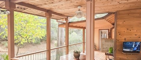 Una terraza de madera con una mesa y sillas, un ventilador de techo y un televisor montado en la pared.