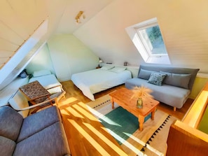 Et kompakt og moderne loftsværelse med skråvæg, seng, sofa, et lille bord og et vindue.