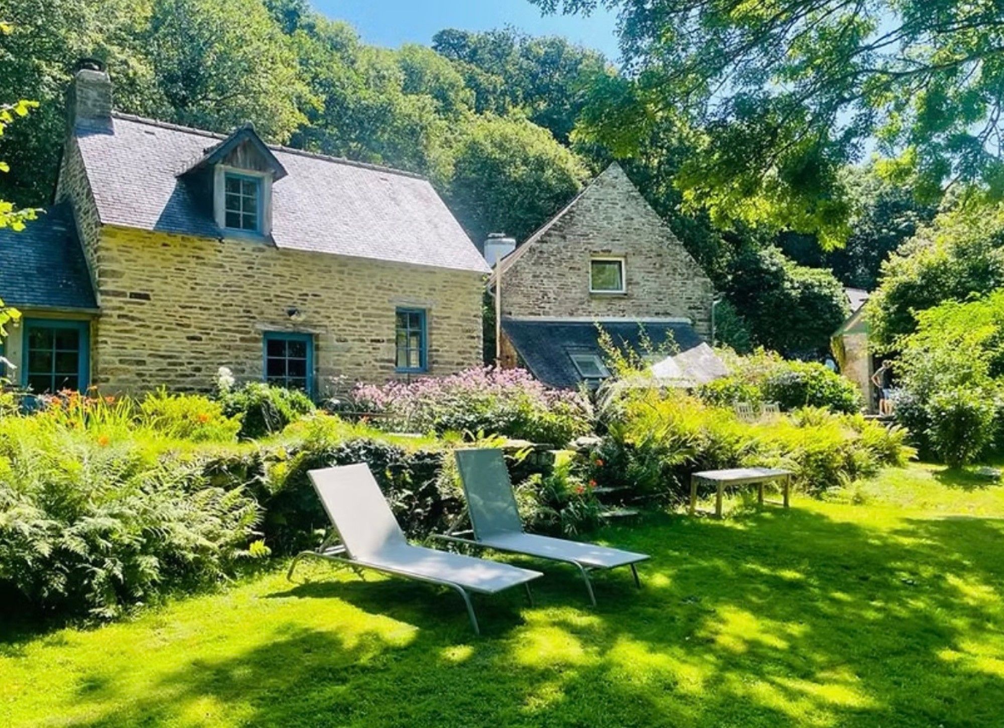 A stone house with a sloped roof and a garden with two lounge chairs.