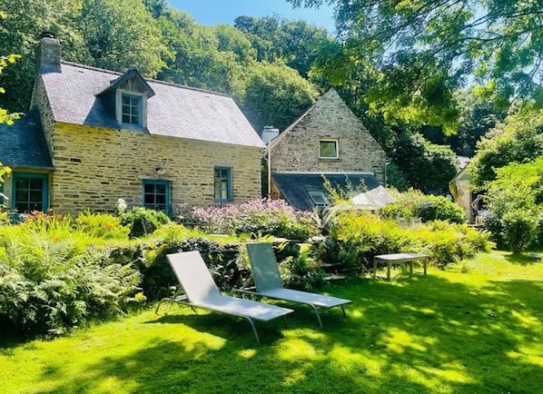 A stone house with a sloped roof and a garden with two lounge chairs.