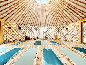 Ein geräumiger Raum mit hoher Decke und einer Wand aus Holzgitter. Mehrere Yogamatten liegen ordentlich auf dem Boden ausgerollt.