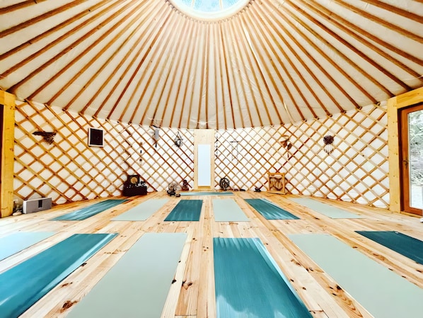 A spacious room with a high ceiling and a wooden lattice wall. There are multiple yoga mats aligned on the floor.