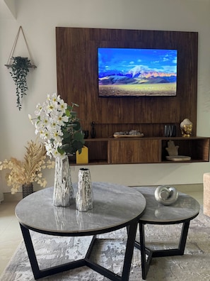 A modern living room with a flat-screen TV mounted on a wooden wall, two round tables with metal frames, and decorative items including vases and a Buddha statue.