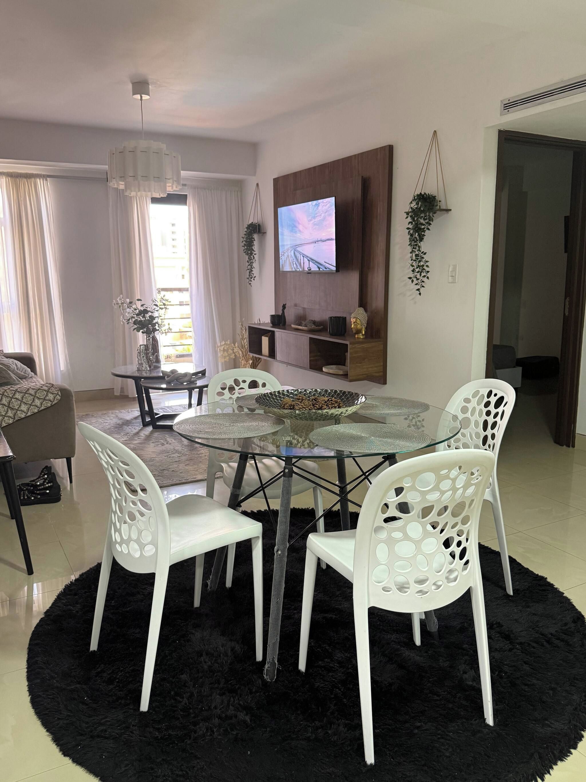 Een moderne woonkamer met een ronde glazen tafel, witte stoelen, een aan de muur bevestigde televisie en een bijzettafeltje met decoratieve objecten.