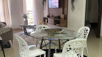 Een moderne woonkamer met een ronde glazen tafel, witte stoelen, een aan de muur bevestigde televisie en een bijzettafeltje met decoratieve objecten.