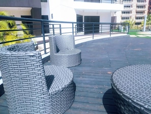 Outdoor wicker furniture on a wooden deck with a railing, overlooking a building with multiple balconies.