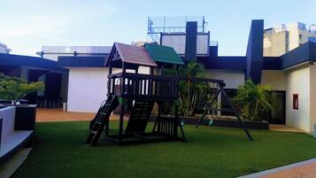 A playground with a slide and swings, surrounded by artificial grass, in front of a modern building with a flat roof.