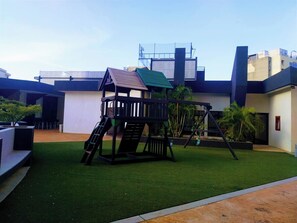 A playground with a slide and swings, surrounded by artificial grass, in front of a modern building with a flat roof.