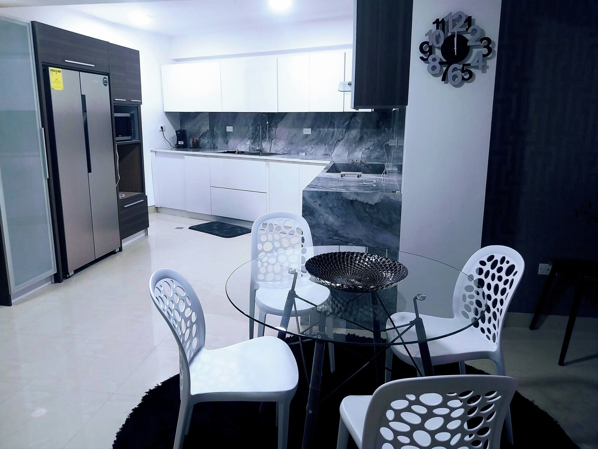 A modern kitchen with white cabinets, a stainless steel refrigerator, and a glass dining table with four chairs.