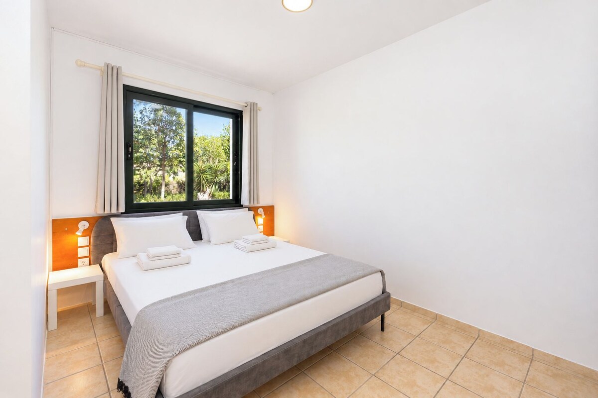 A hotel room with a bed, bedside tables, a window with curtains, and a view of greenery outside.