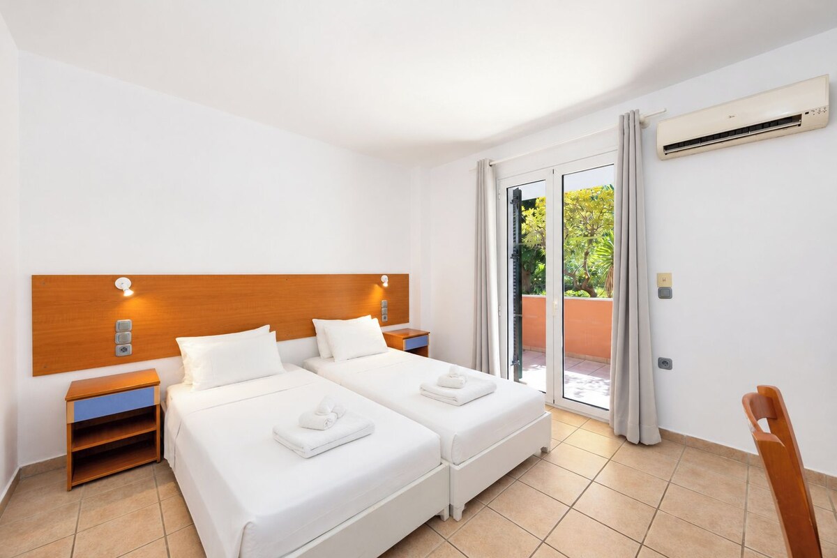 A hotel room with two beds, a wooden headboard, a nightstand, a chair, and a sliding glass door leading to an outdoor area.