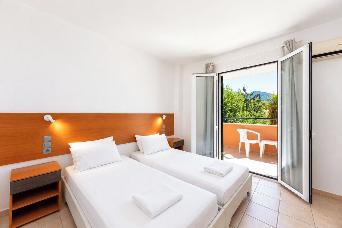 A hotel room with two beds, a nightstand, a window with a view of greenery, and a sliding glass door to a balcony.