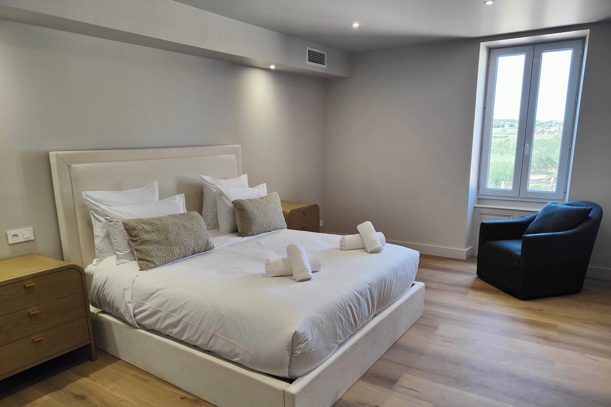 A modern bedroom with a large bed, a nightstand, a chair, and a window with a view.