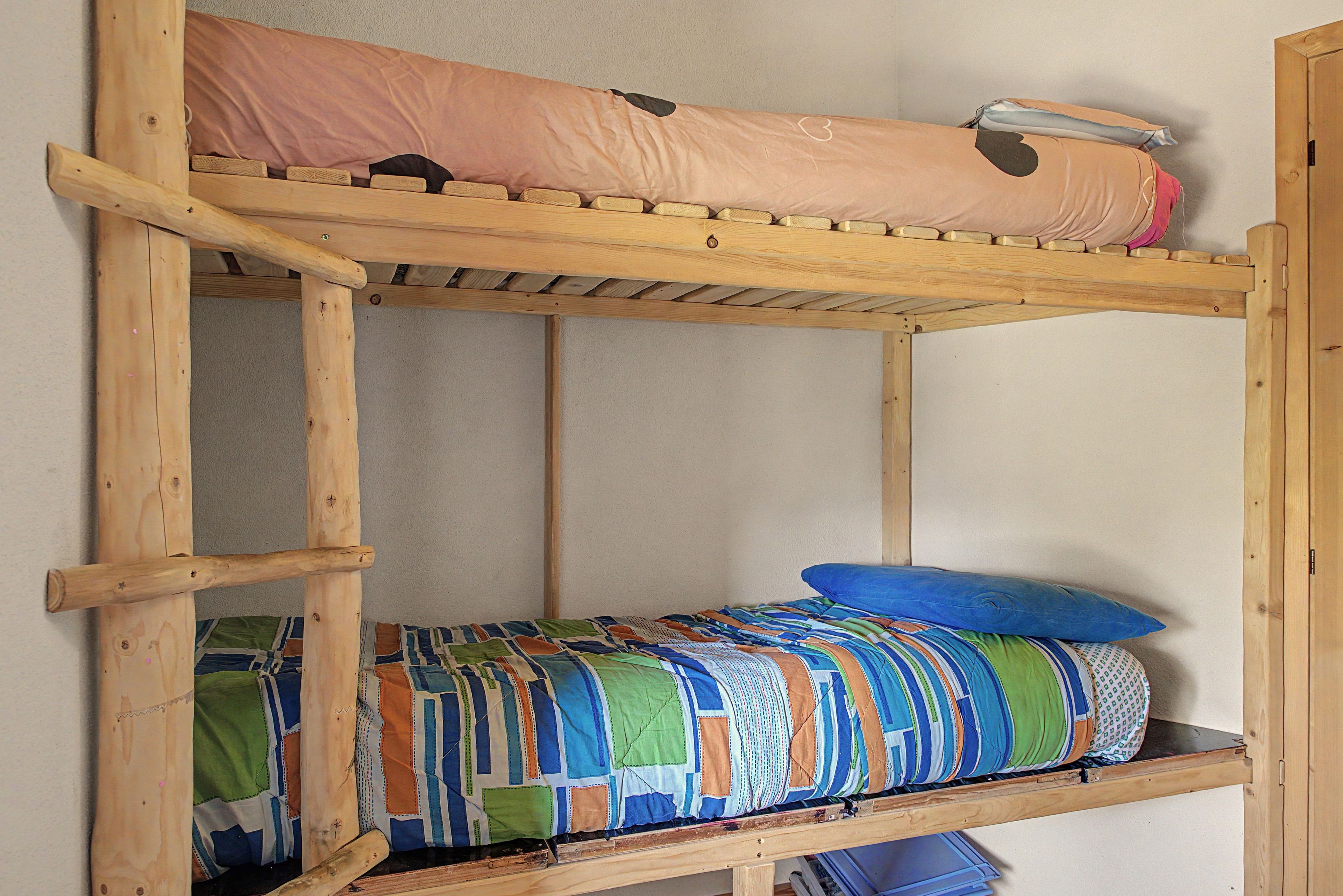 Een stapelbed met houten frame, een bovenbed met een roze matras en een onderbed met een kleurrijke quilt.