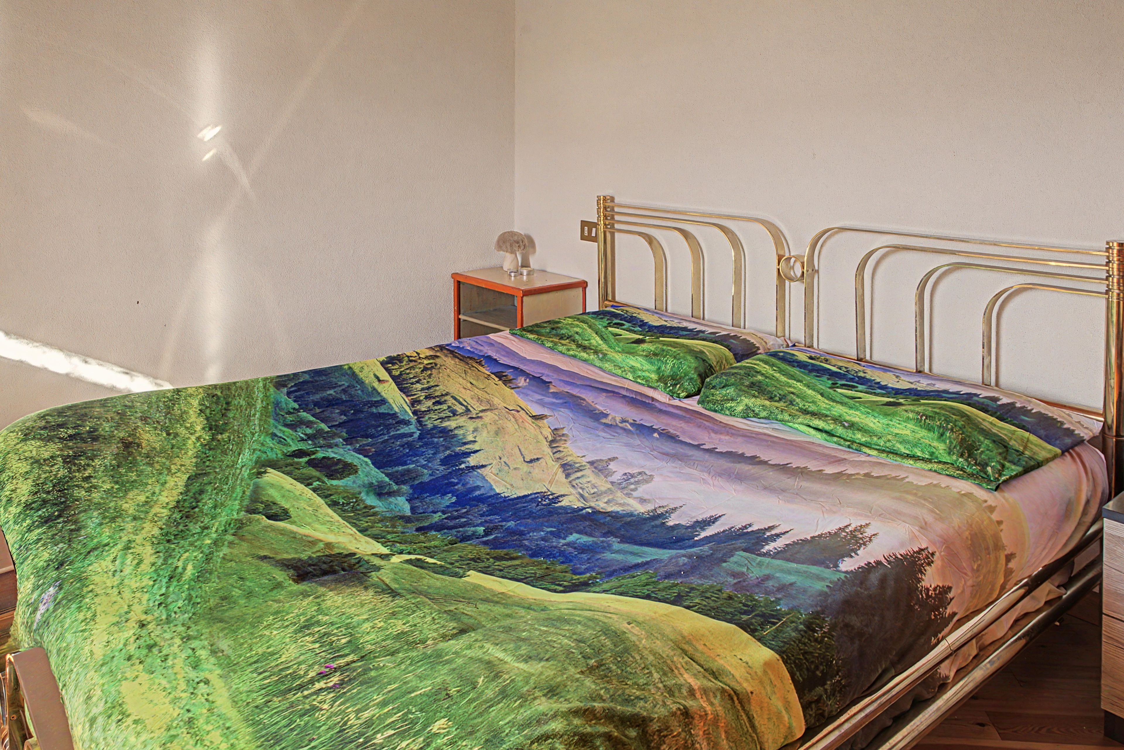 Una cama con estructura metálica y una colcha con motivos de paisaje.