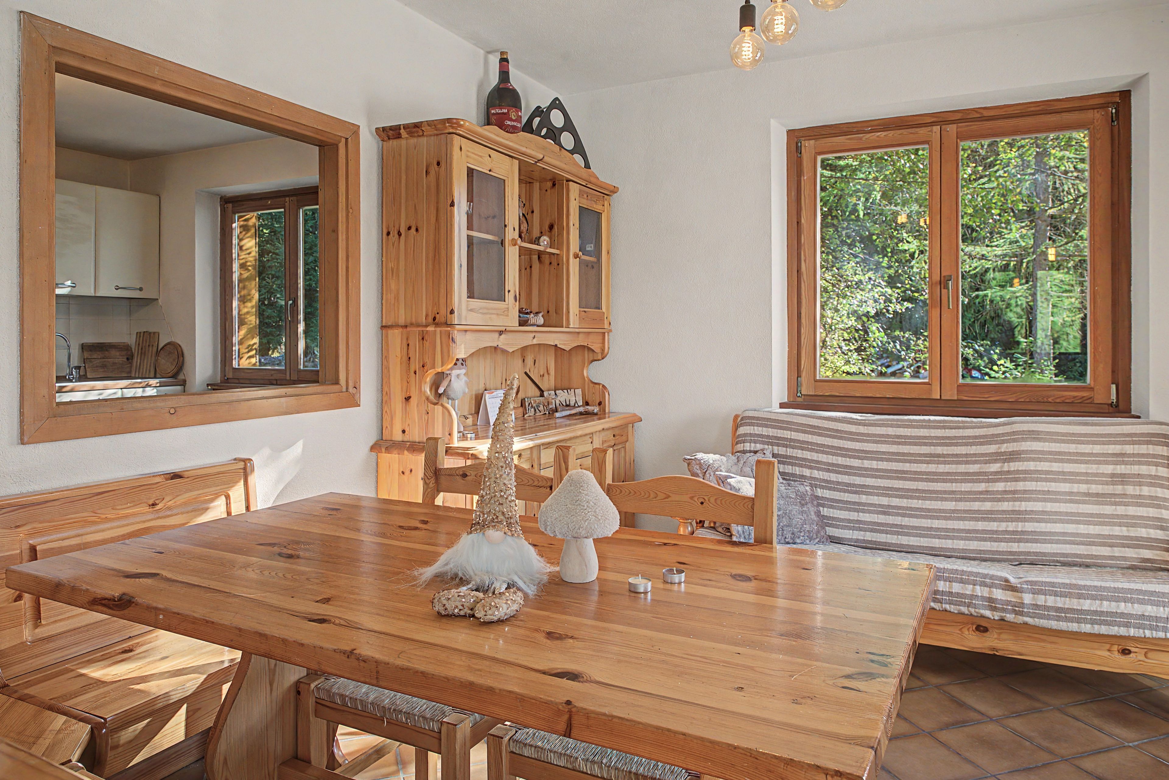 A wooden dining table with benches, a wooden cabinet with glass doors, a gnome decoration, and a window with a view of trees.
