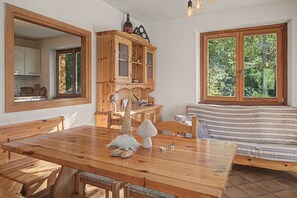 Een houten eettafel met banken, een houten kast met glazen deuren, een tuinbeeldje en een raam met uitzicht op bomen.
