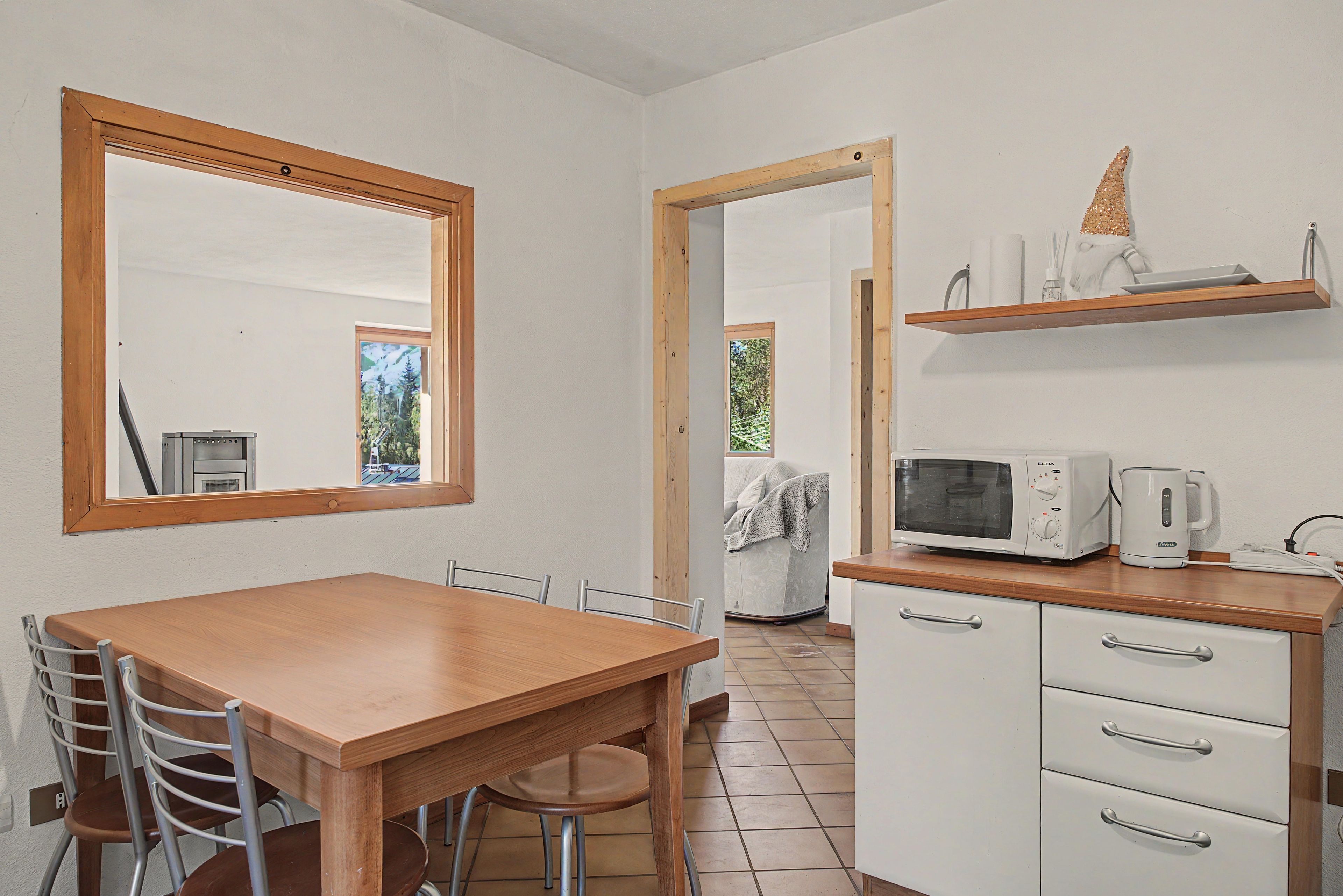 Een keuken met een houten tafel, stoelen, een magnetron, een waterkoker en een plank met keukenspullen.