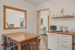 A kitchen with a wooden table, chairs, a microwave, a kettle, and a shelf with kitchen items.
