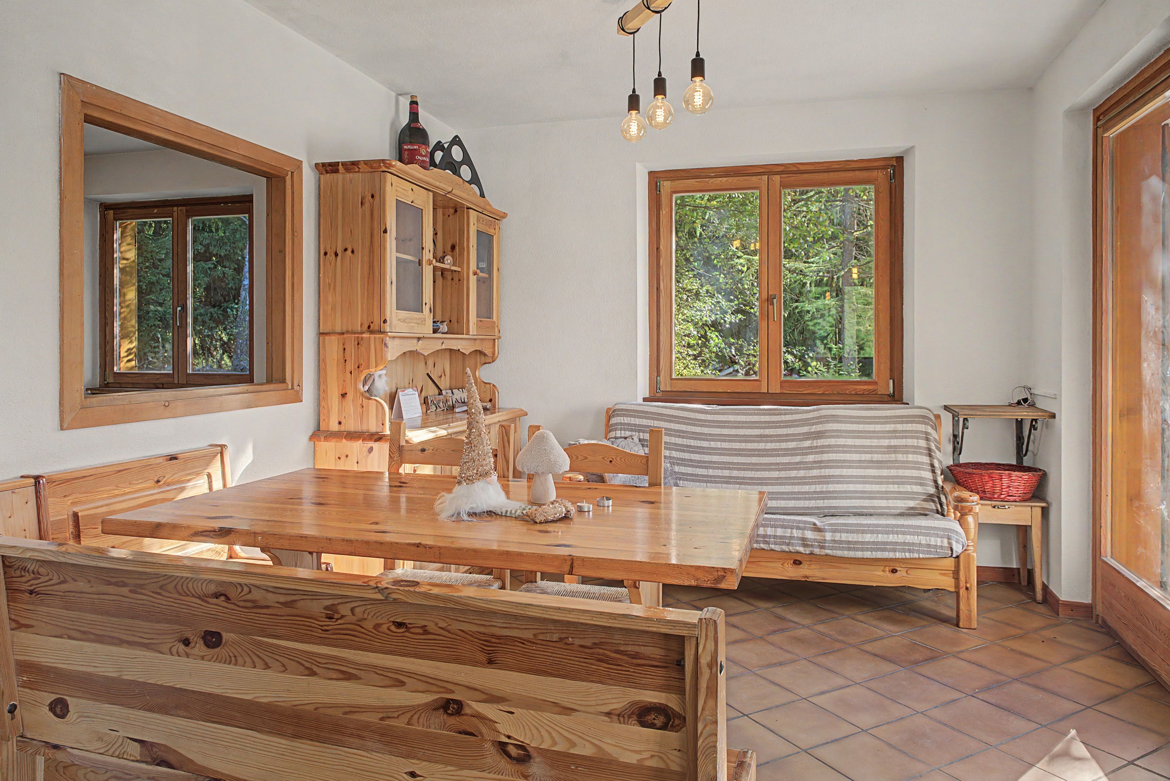 A wooden dining table with chairs, a wooden cabinet, a bed with a striped blanket, and a window with a view of trees.