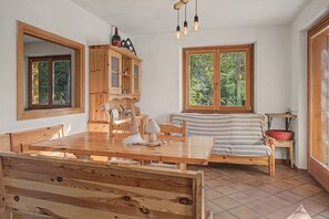 Una mesa de comedor de madera con sillas, un armario de madera, una cama con manta a rayas y un ventanal con vistas a árboles.