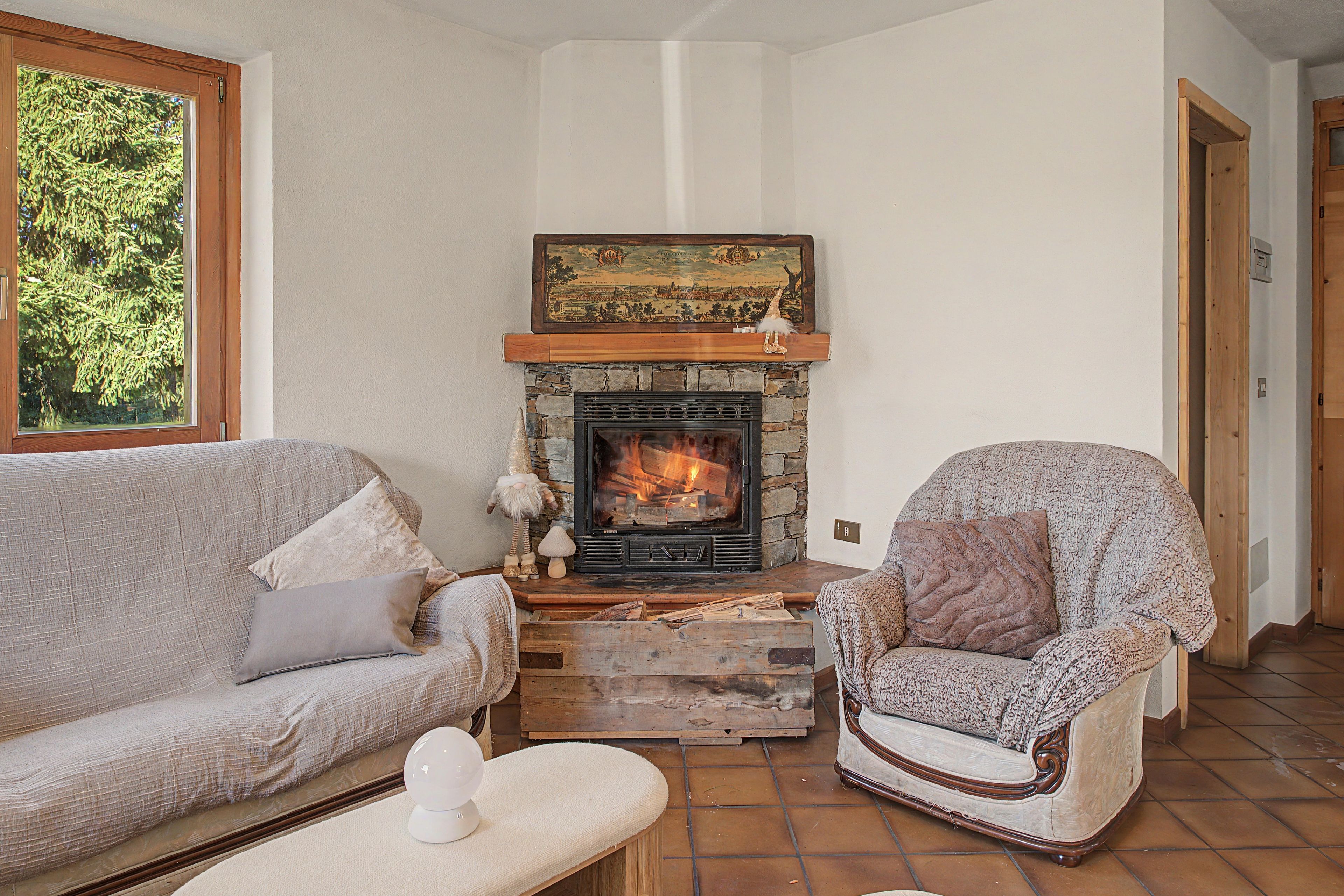 Een gezellige woonkamer met een stenen open haard, een bank, een fauteuil met een patroon en een raam met uitzicht op bomen.