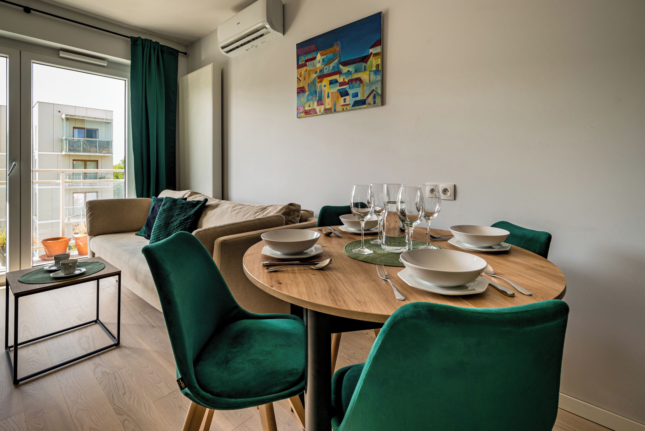 Un comedor moderno con una mesa redonda de madera, sillas tapizadas en verde y vistas a un balcón con un paisaje urbano.