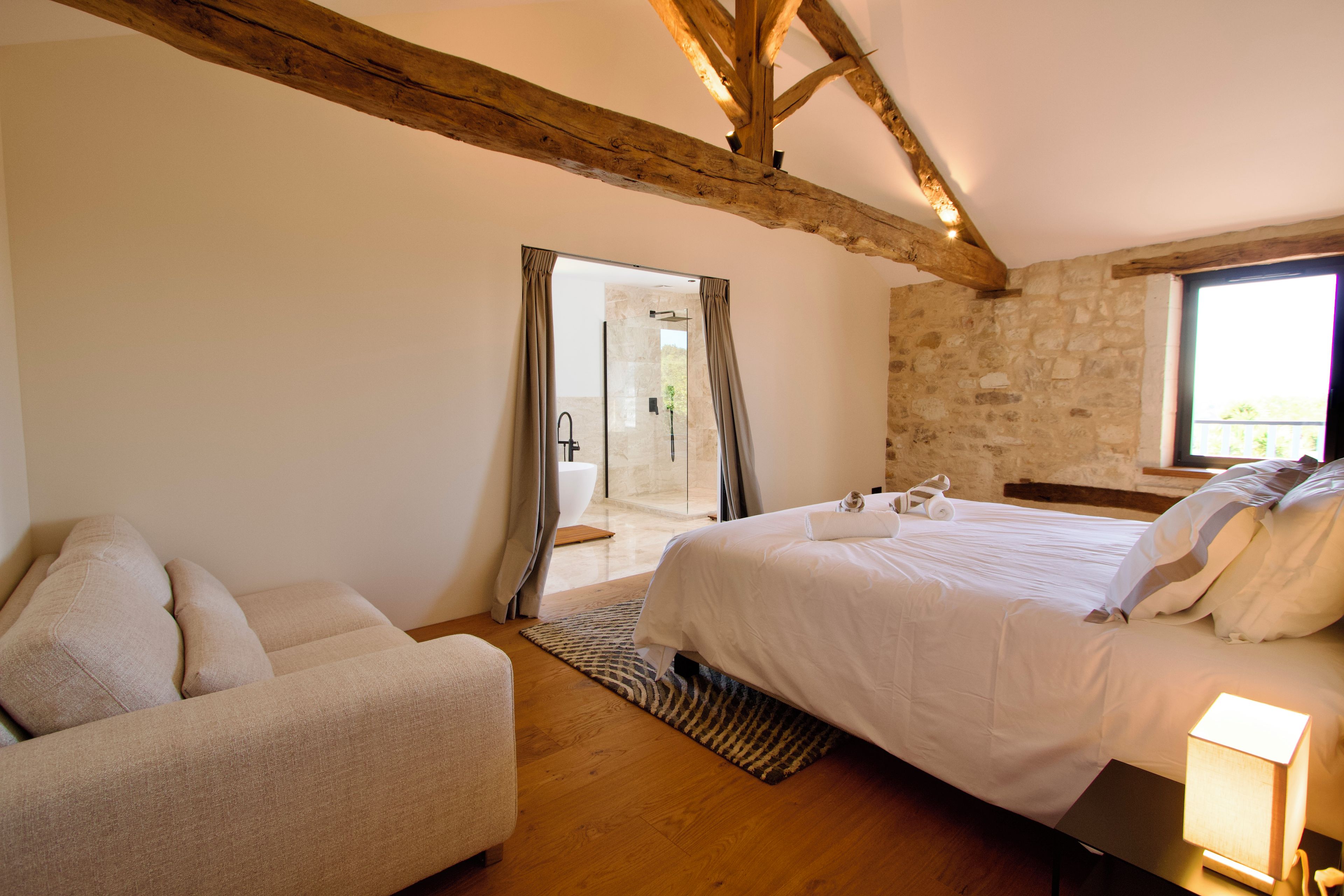 Un dormitorio con cama, una silla, mesita de noche con lámpara, zona de ducha y pared de piedra.