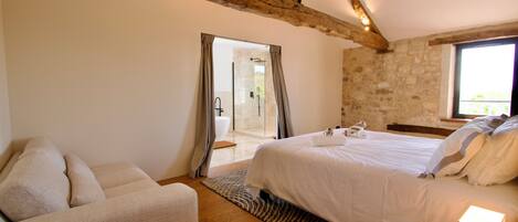 Un dormitorio con cama, una silla, mesita de noche con lámpara, zona de ducha y pared de piedra.