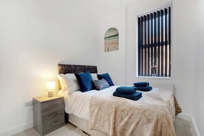 A bedroom with a bed, bedside table, lamp, and a window with blinds. - Ground-Floor 2BR Newcastle Retreat | Sleeps 4 (Newcastle upon Tyne)