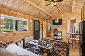 L’intérieur d’un chalet en bois comprend un lit, une table avec des magazines, un téléviseur et une fenêtre donnant sur une forêt.