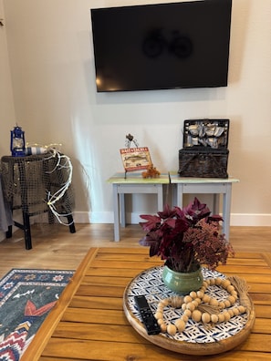 Een woonkamer met een aan de muur bevestigde televisie, een rieten fauteuil, een tafeltje met een bord, een mand, een vaas met bloemen en een afstandsbediening op een dienblad.