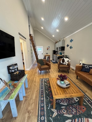 Een moderne woonkamer met een houten salontafel, twee bruine fauteuils, een bank, een aan de muur bevestigde televisie en een trap naar de bovenverdieping.