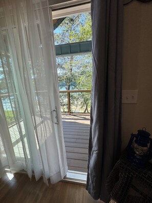 Ein Zimmer mit einer Holterrasse, Blick auf Bäume und einen klaren Himmel sowie einer Tür mit weißen Vorhängen.