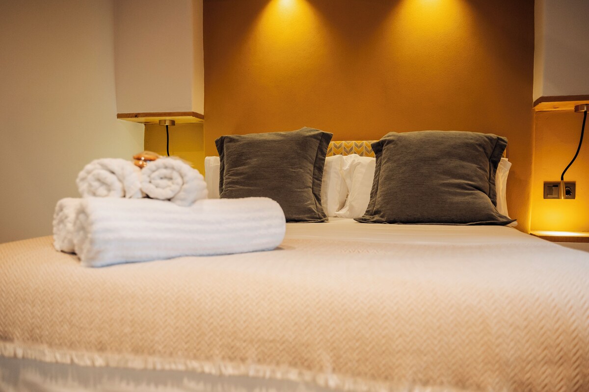 A neatly made bed with pillows and rolled towels, a bedside lamp, and a wall-mounted light fixture.