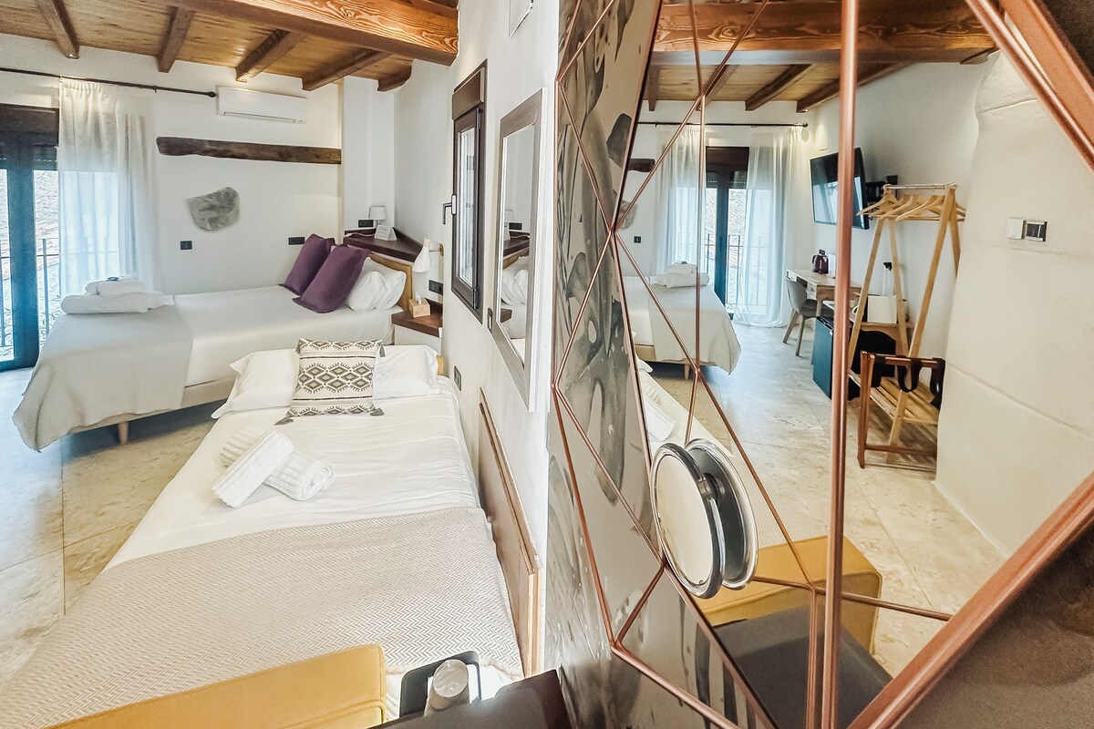 A hotel room with a bed, a desk, and a chair. There is a window with curtains, a mirror, and a towel rack.