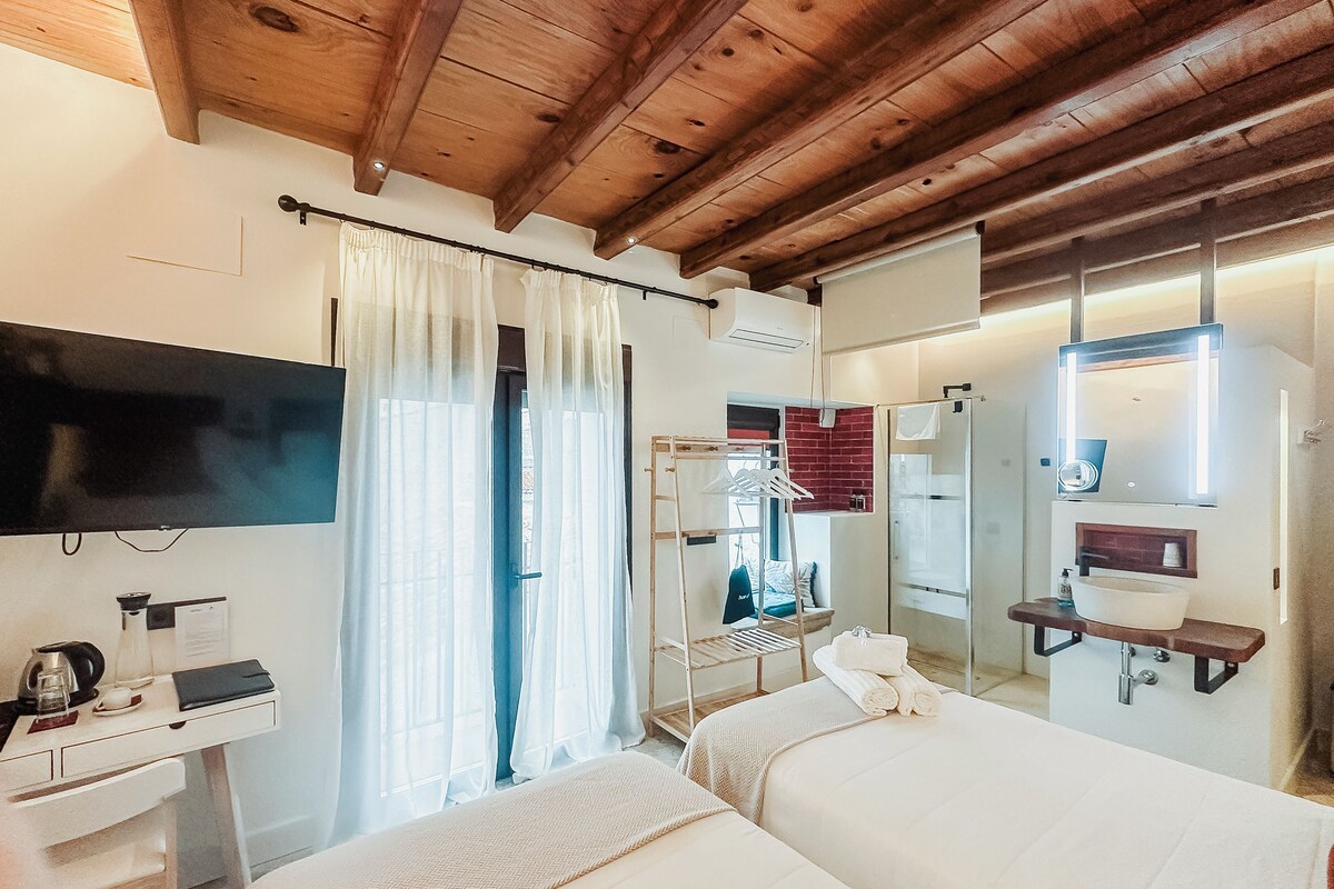 A hotel room with a wooden ceiling, a bed, a desk with a telephone, a mirror, and a window with curtains.